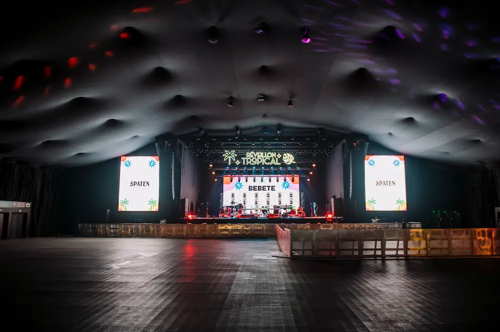 Arena fechada com o palco principal, telões e estrutura de iluminação da edição anterior.